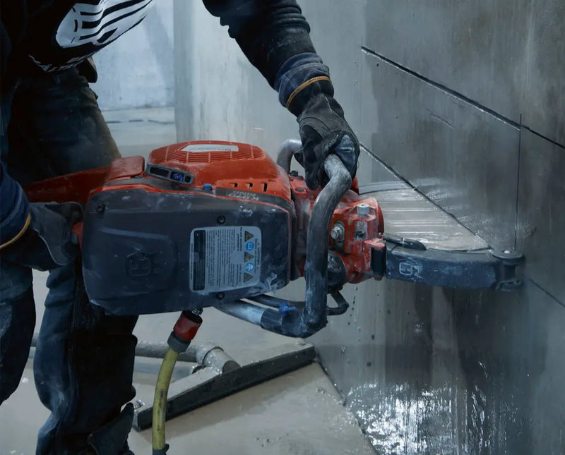 Construction worker in protective clothing, gloves, and safety equipment utilizing a high-powered orange ring saw to cut horizontal lines into a concrete wall.

